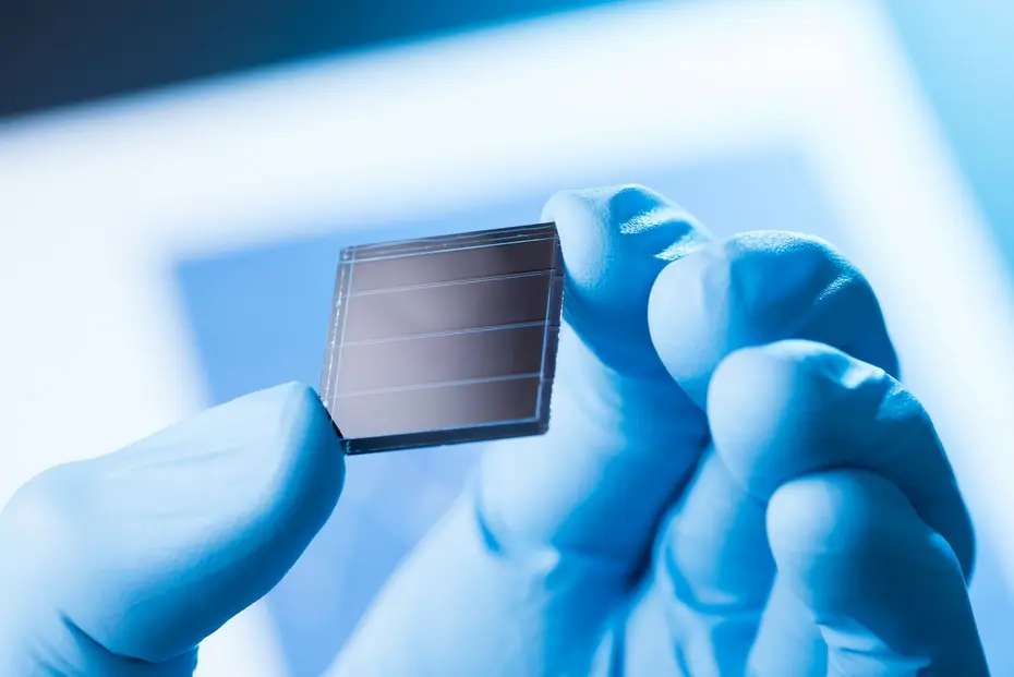Laborhand mit Handschuh haelt eine kleinformatige Solarzelle mit sichtbaren Kontaktstreifen vor blauem Hintergrund