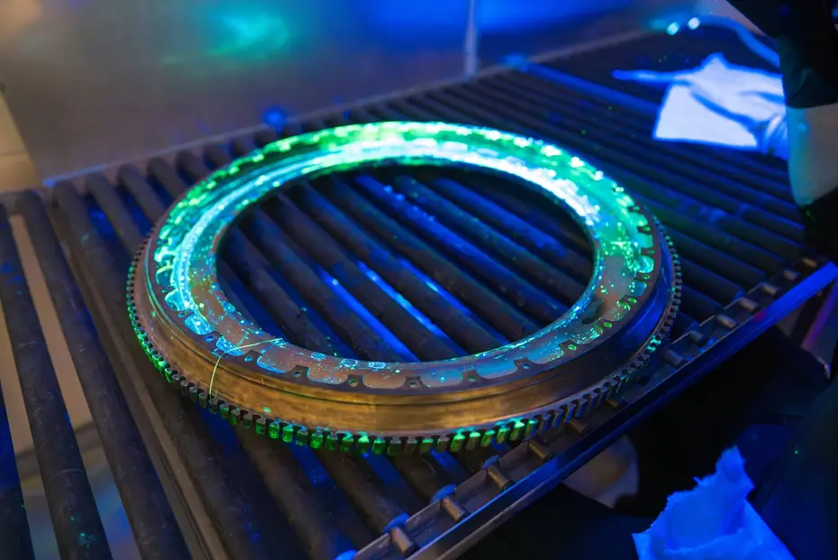 Ringförmiges, verzahntes Metallbauteil in einer dunklen Prüfkabine mit grün fluoreszierenden Eindringmittel-Anzeigen entlang der Zahnflanken unter UV-A-Bestrahlung, Prüfperson mit Tuch im Hintergrund
